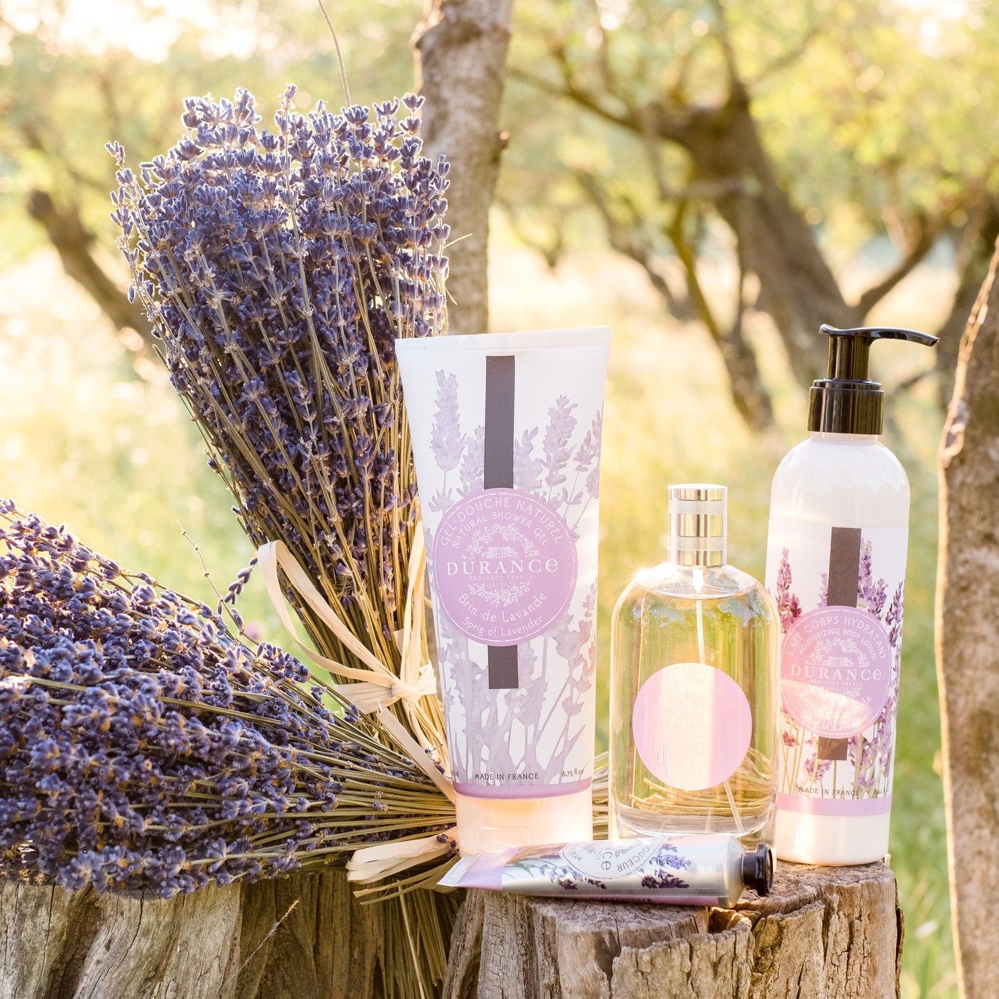 Lavender-themed products including a bottle, tube, and pump on a wooden stump with lavender flowers.Sprig of Lavender - Shower Gel