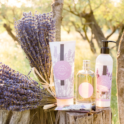 Lavender-themed products including a bottle, tube, and pump on a wooden stump with lavender flowers.Sprig of Lavender - Shower Gel 