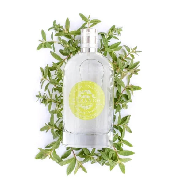 A bottle of Durance sparkling verbena eau de toilette perfume next to its packaging box, both featuring a green label with images of verbena leaves 